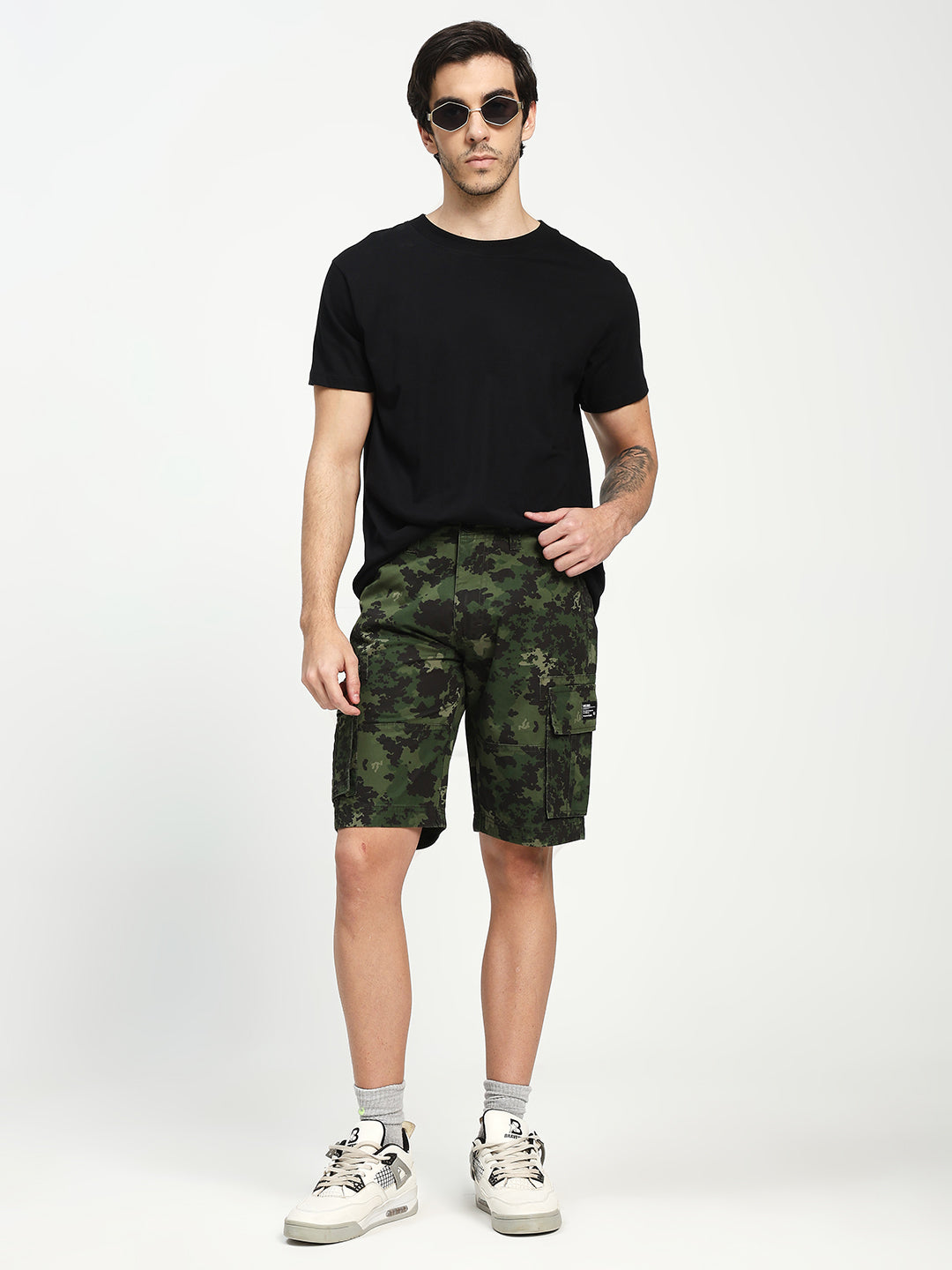 Man wearing a black t-shirt, green camouflage shorts, and white sneakers on a white background