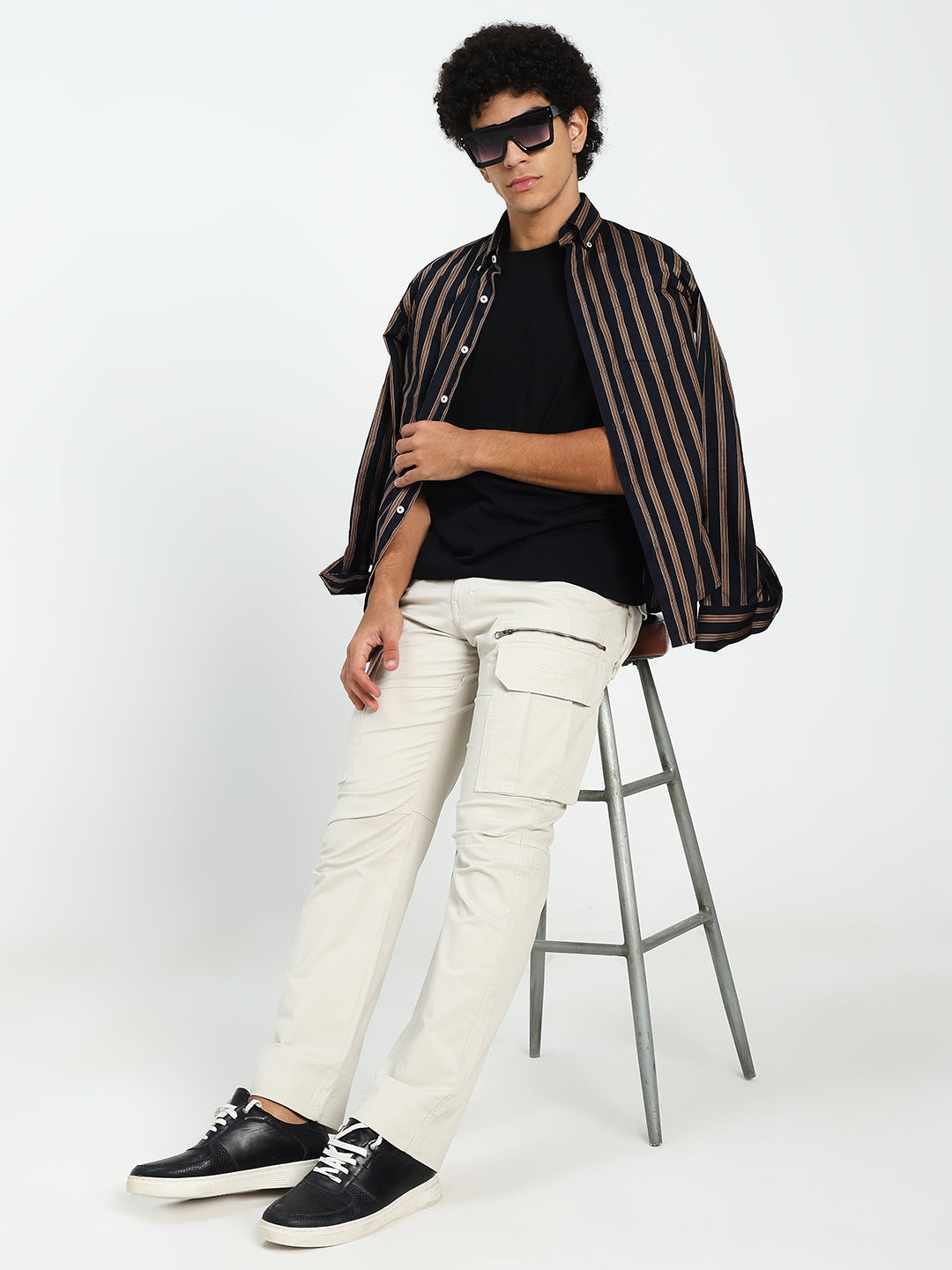 man sitting on a stool wearing a striped shirt, black top, cream cargo pants, and black shoes on a white background