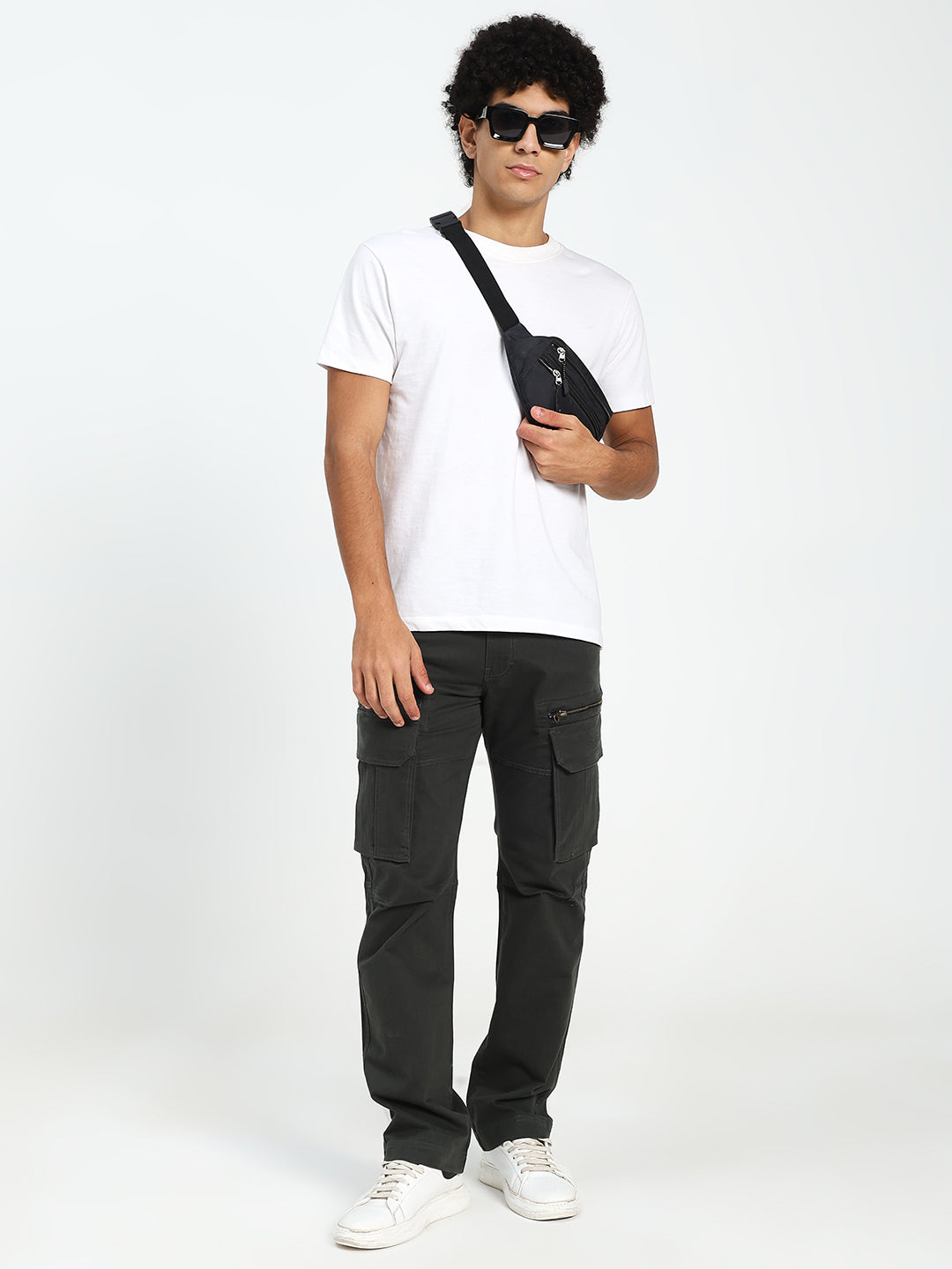 Man wearing a white t-shirt, dark green cargo pants, and sunglasses holding a black bag on a white background