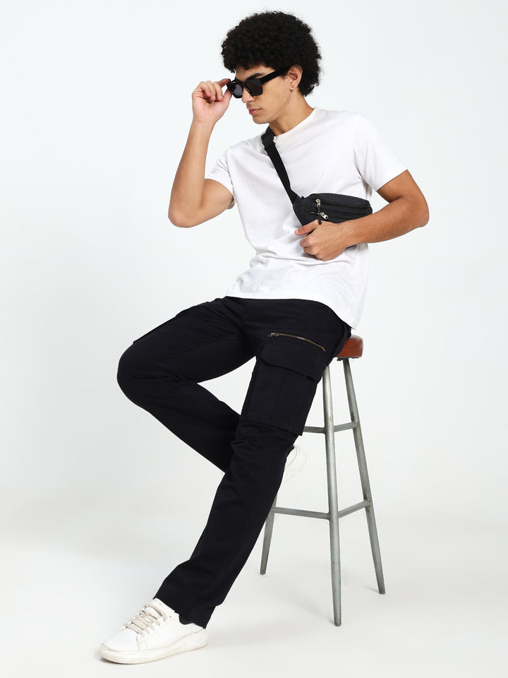 man sitting on a stool wearing a white t-shirt, black cargo pants, and sunglasses with a black bag over one shoulder.