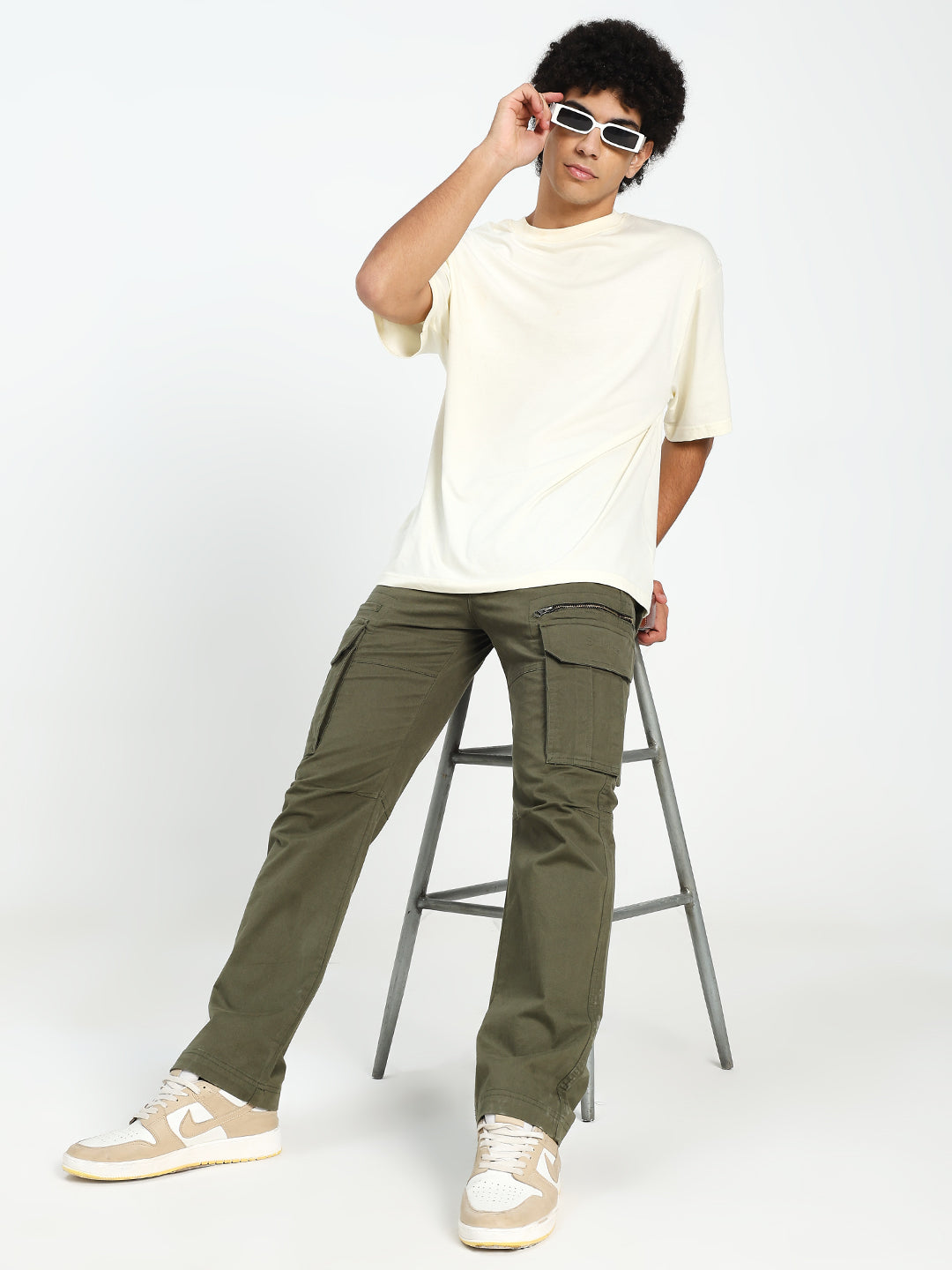 man wearing a white t-shirt and olive green cargo pants sitting on a stool against a white background
