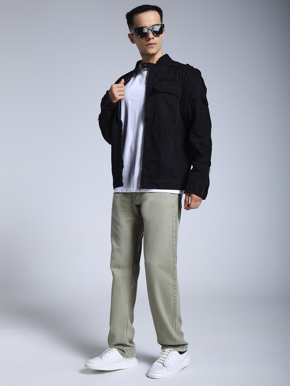 Man wearing a black jacket, white shirt, green pants, and white sneakers on a gray background