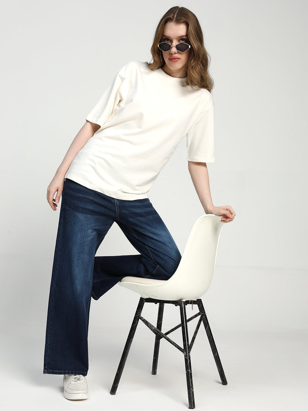 Woman sitting on a white chair wearing a white t-shirt and blue jeans against a plain background