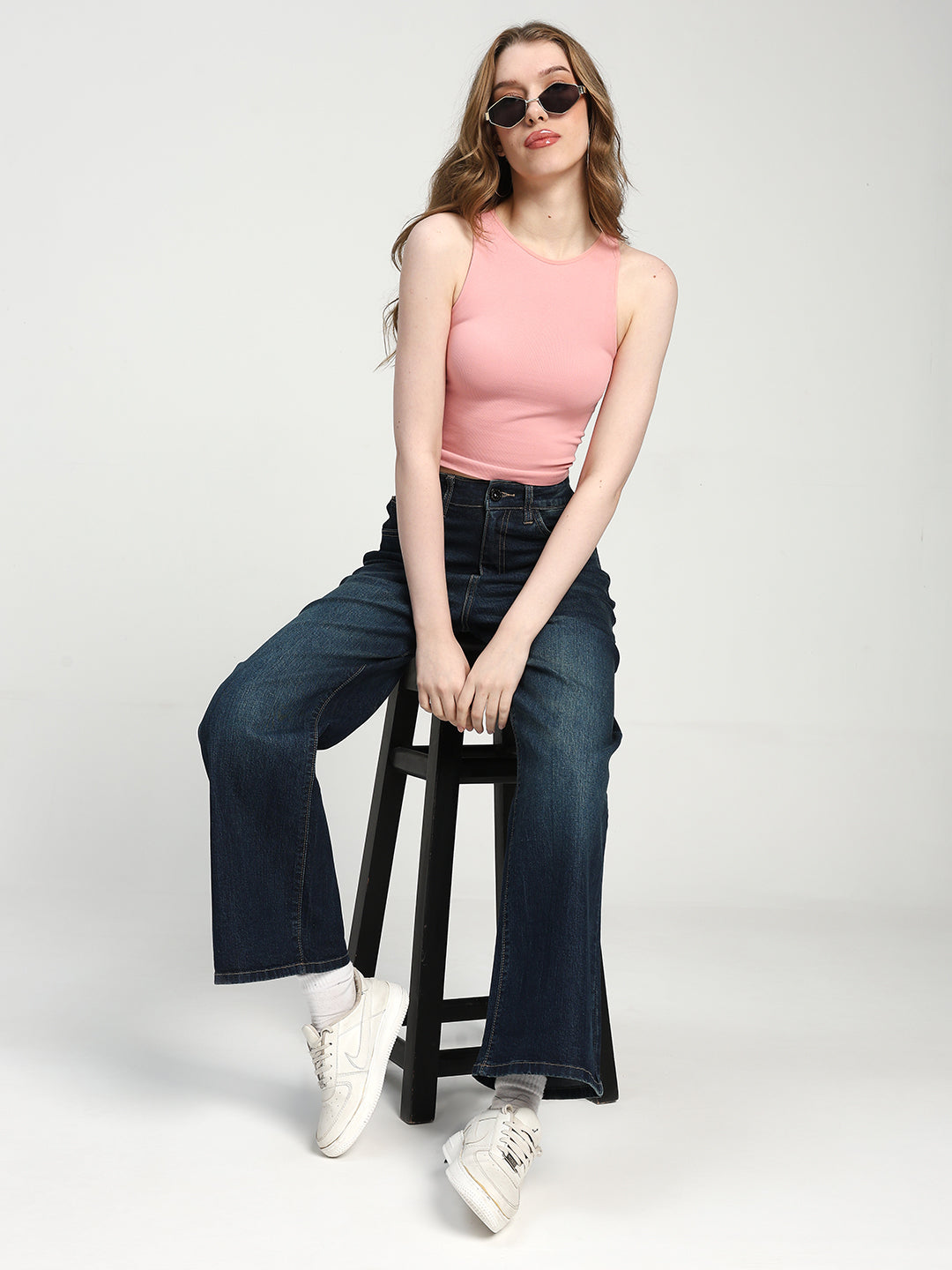 Woman sitting on a stool wearing a pink sleeveless top and dark blue jeans against a white background