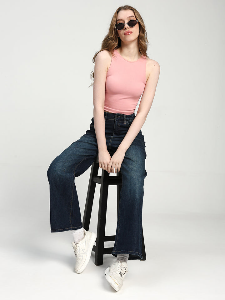 Woman sitting on a stool wearing a pink sleeveless top and dark blue jeans against a white background