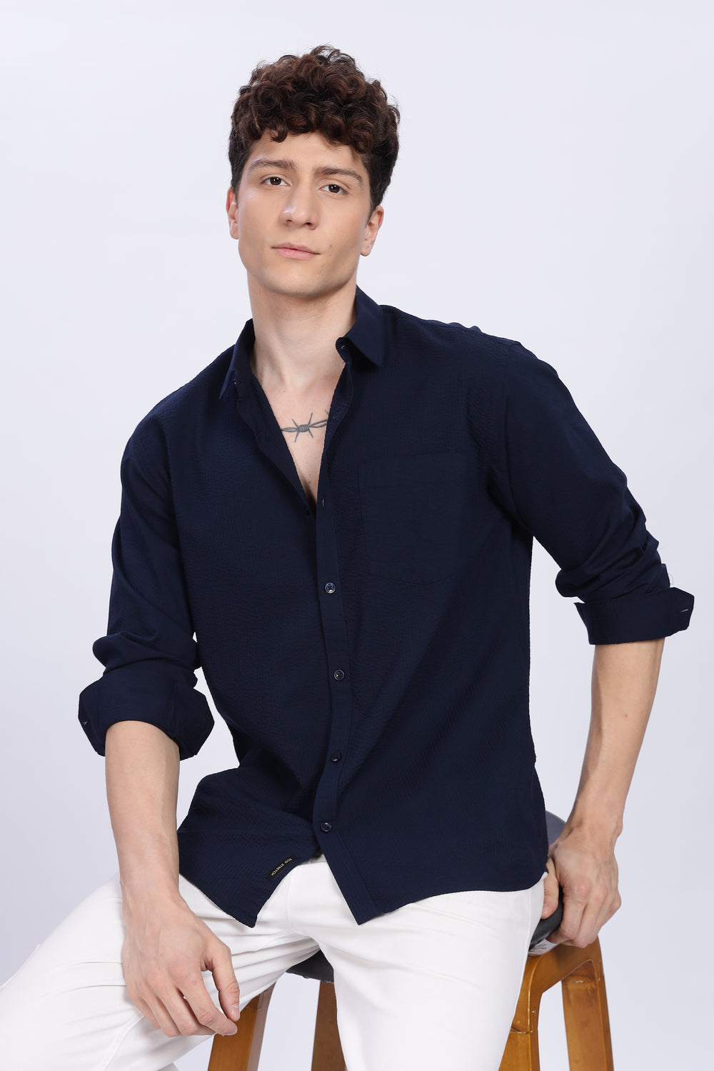Man wearing a navy blue shirt and white pants sitting on a wooden stool against a white background