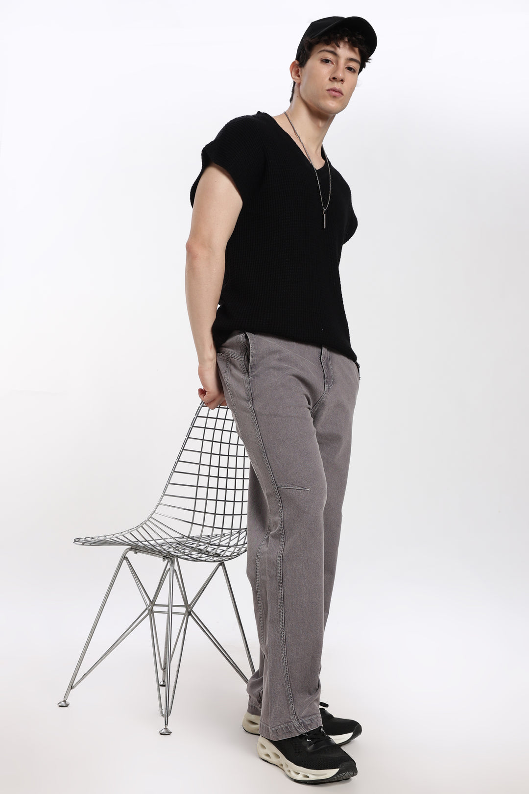 man wearing a black sleeveless t-shirt and gray jogger standing next to a wire chair on a white background