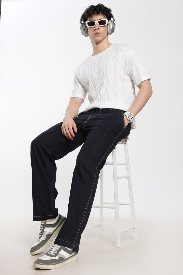 man wearing a white shirt, blue jogger, and sunglasses sitting on a stool against a white background