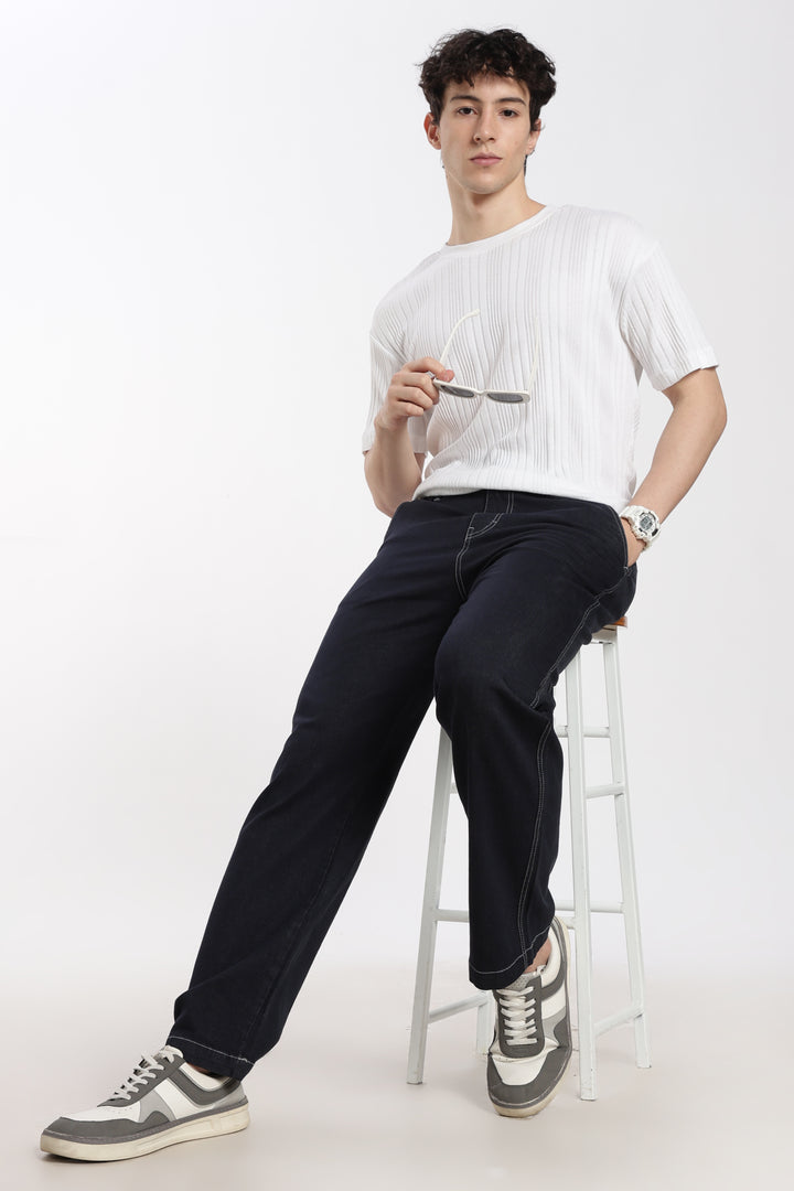 man sitting on a stool wearing a white t-shirt and blue jogger against a white background