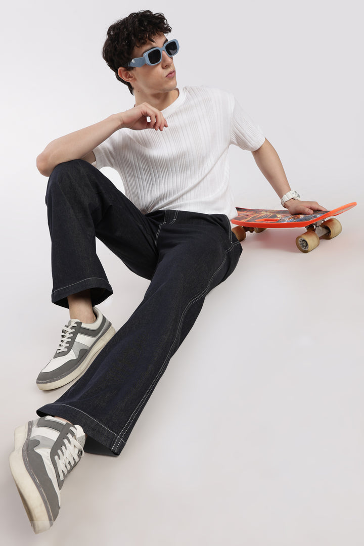 man sitting on a white floor holding a red skateboard, wearing a white shirt, dark wash denim jogger pants, and sunglasses.