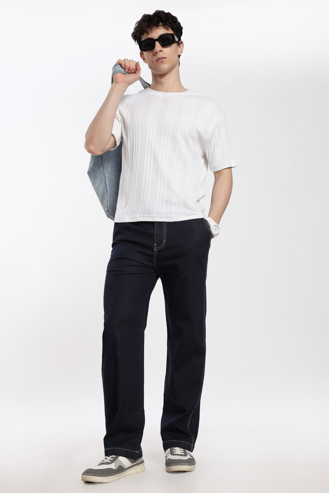 man wearing a white t-shirt and navy blue denim jogger pants holding a blue bag on a white background
