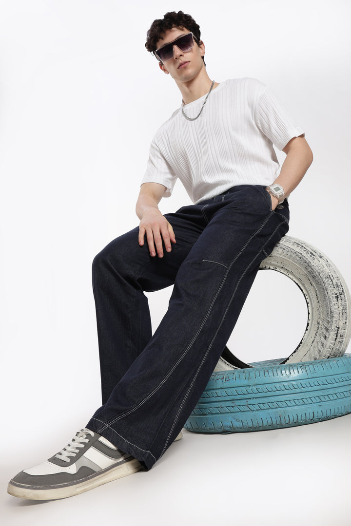 Man sitting on a tire wearing a white shirt, blue denim jogger, and sunglasses on a white background
