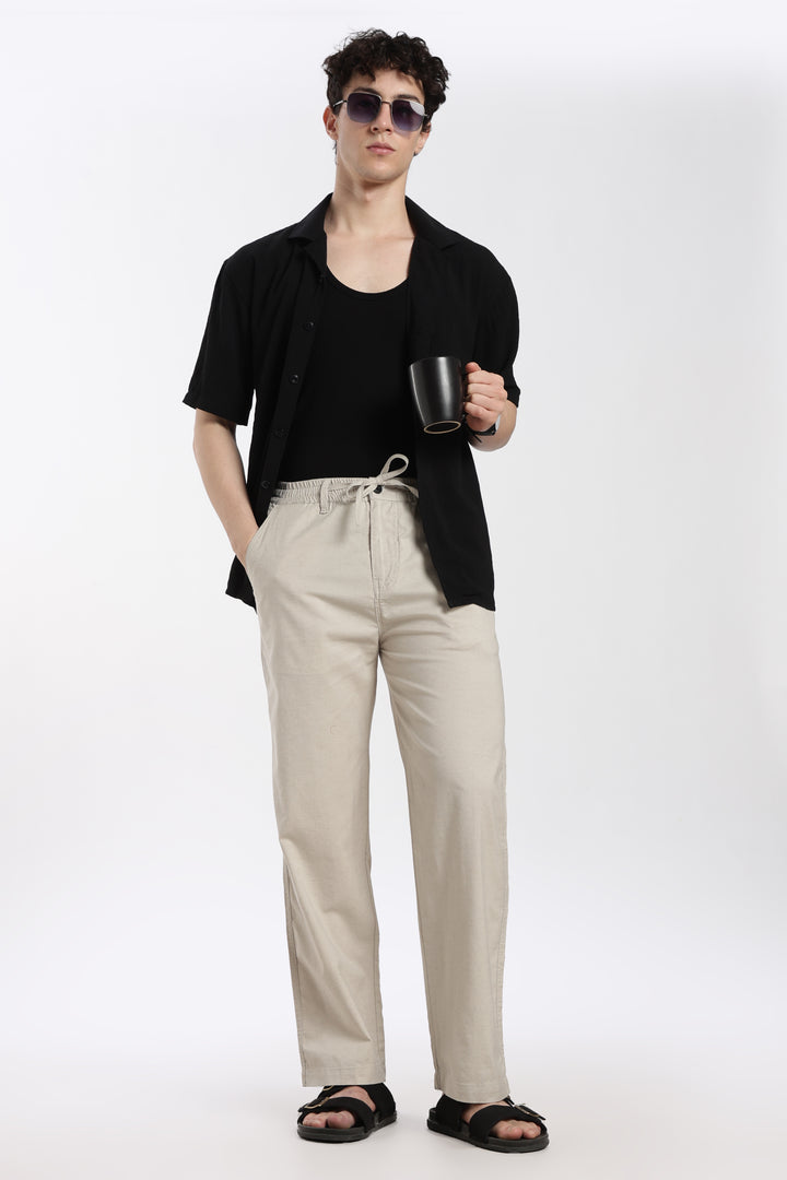 Man wearing a black shirt, desert sand linen pants, and sunglasses holding a mug on a white background
