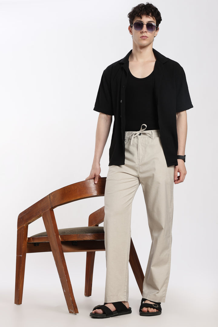 Man wearing a black shirt and desert sand linen pants standing next to a wooden chair on a white background