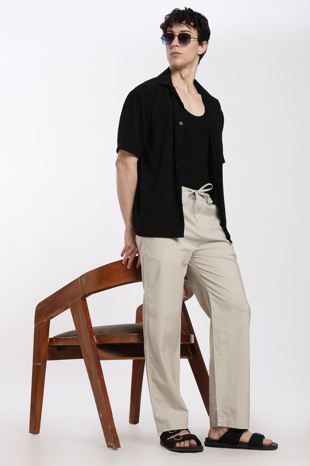 Man wearing a black shirt and desert sand linen pants standing next to a wooden chair on a white background