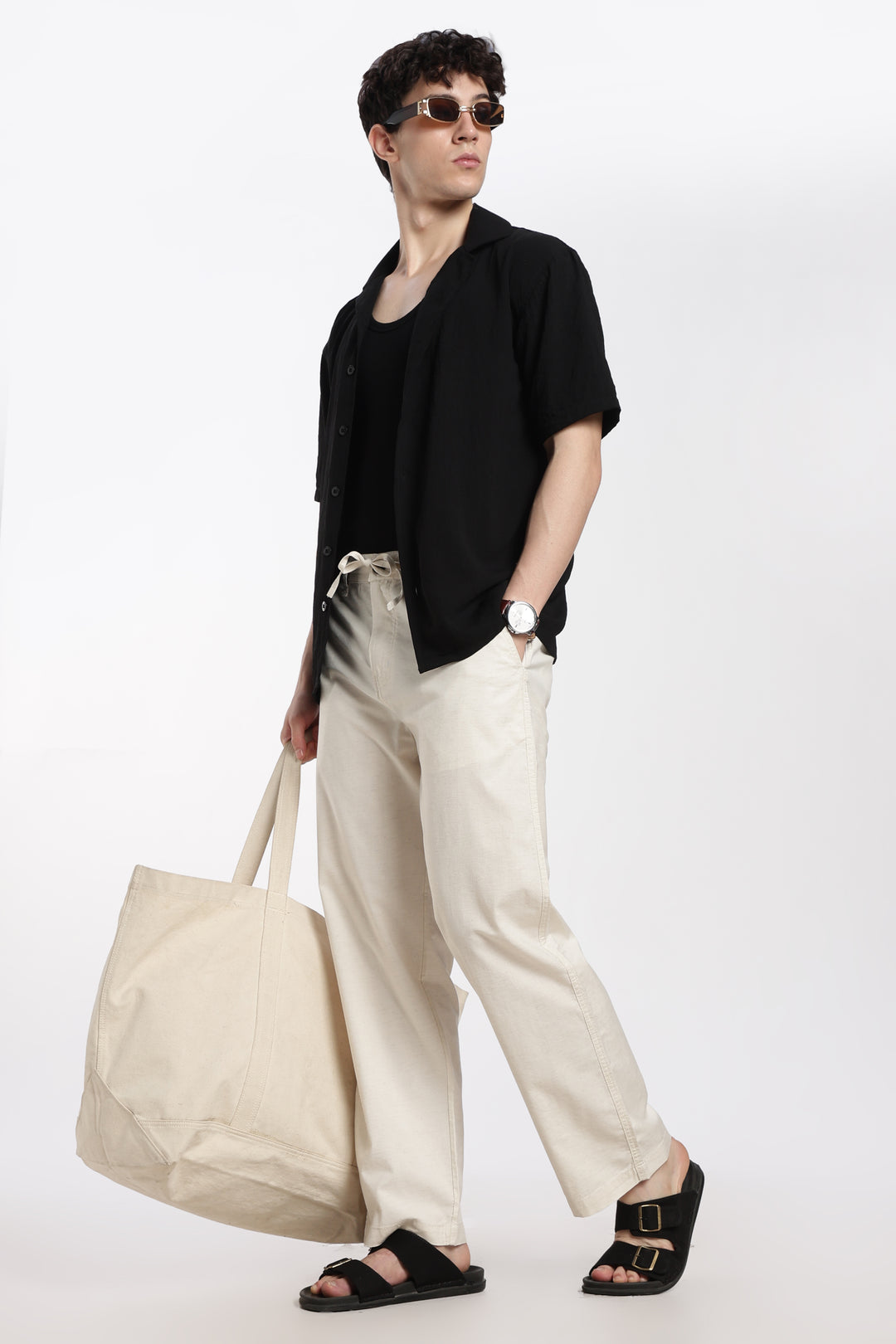man wearing a black shirt and cream linen pants, holding a beige tote bag on a white background