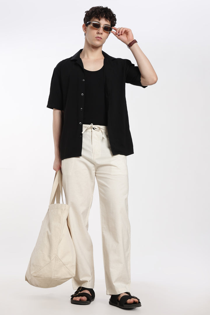 man wearing a black jacket, black shirt, cream linen pants, and sandals, holding a beige bag on a white background.