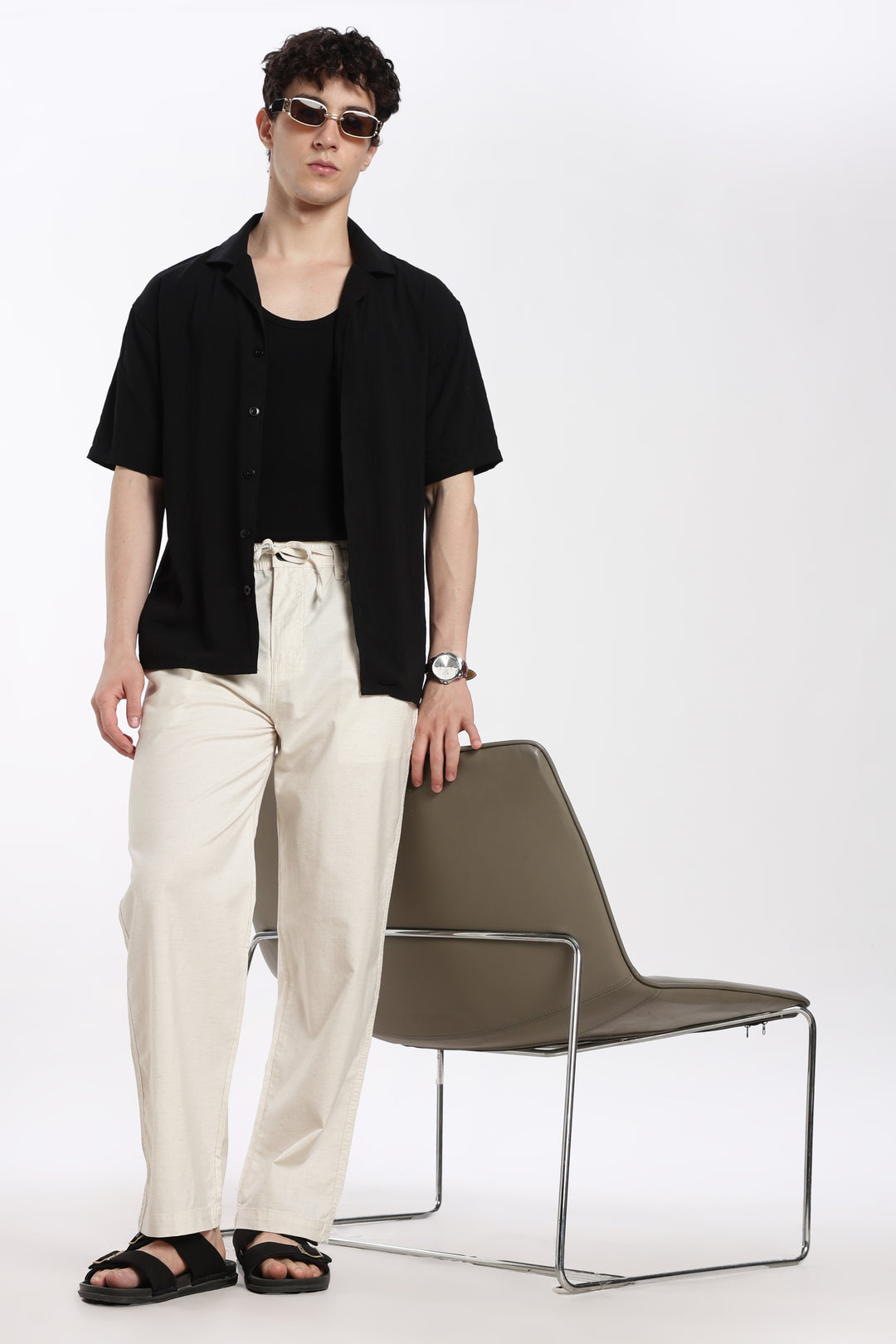 Man wearing a black shirt and cream linen pants standing next to a chair on a white background