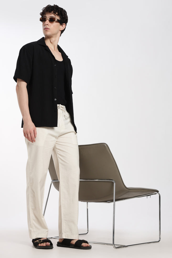 man wearing a black shirt and cream linen pants standing next to a modern chair on a white background