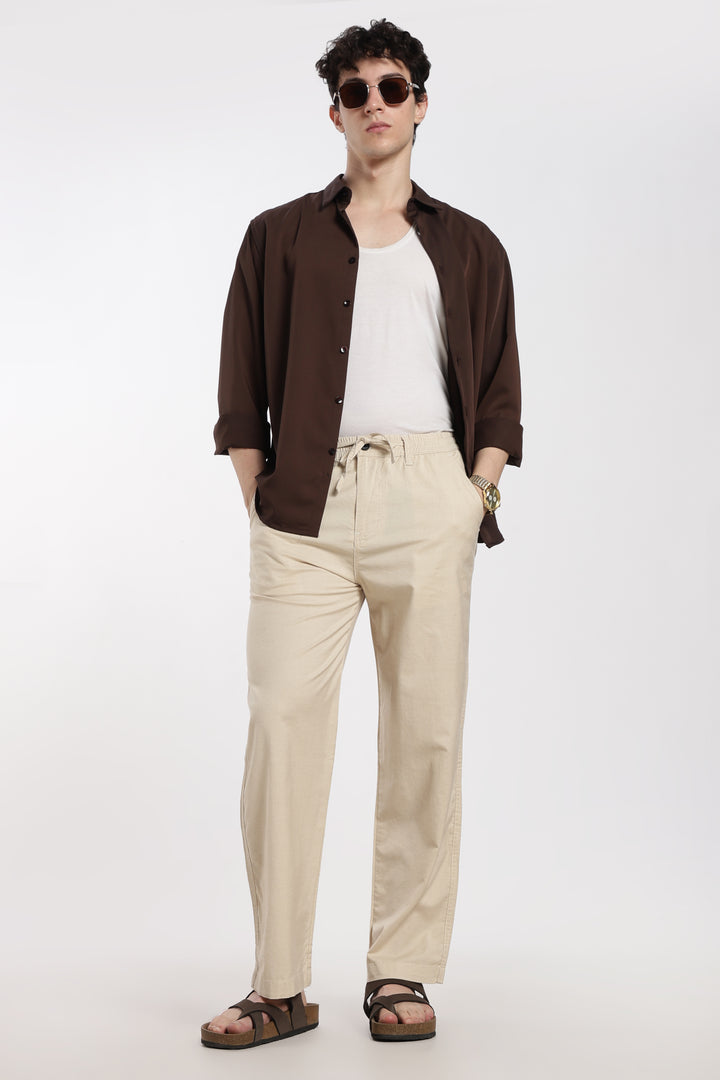 Man wearing a brown shirt, white t-shirt, beige linen pants, and sandals on a white background
