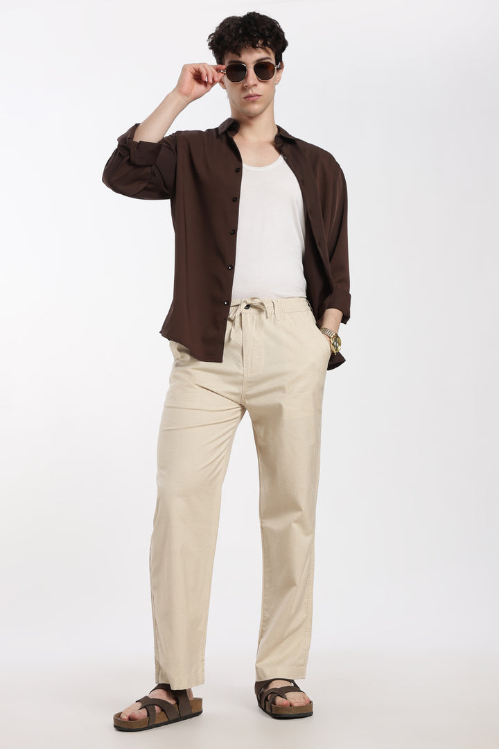Man wearing a brown jacket, white shirt, beige linen pants, and sandals on a white background