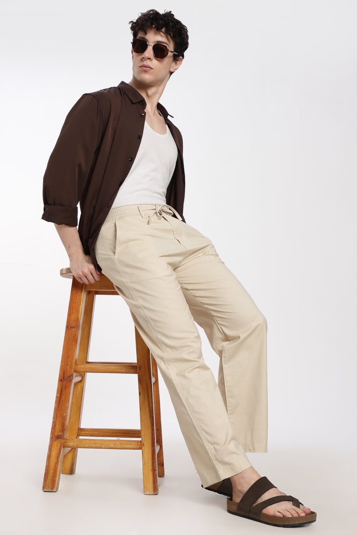 Man sitting on a wooden stool wearing a brown shirt, white tank top, beige linen pants, and sunglasses.