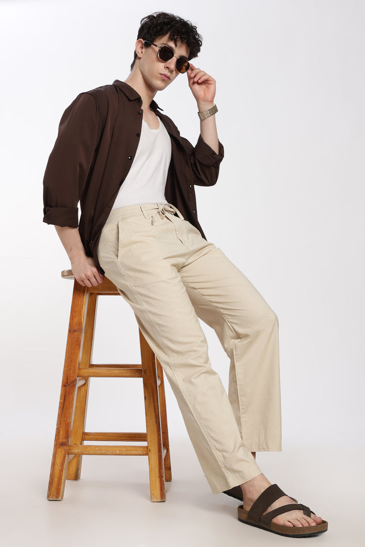 Man sitting on a wooden stool wearing a brown shirt, white tank top, beige linen pants, and sandals.