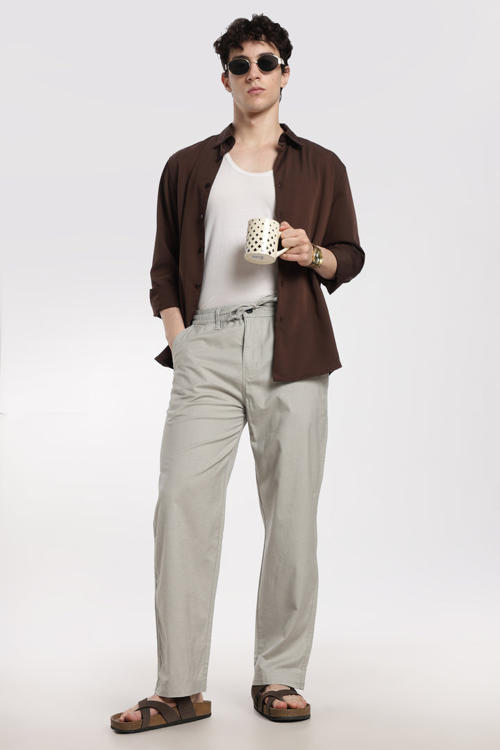 Man wearing a brown shirt, white tank top, grey linen pants, and sandals holding a mug.