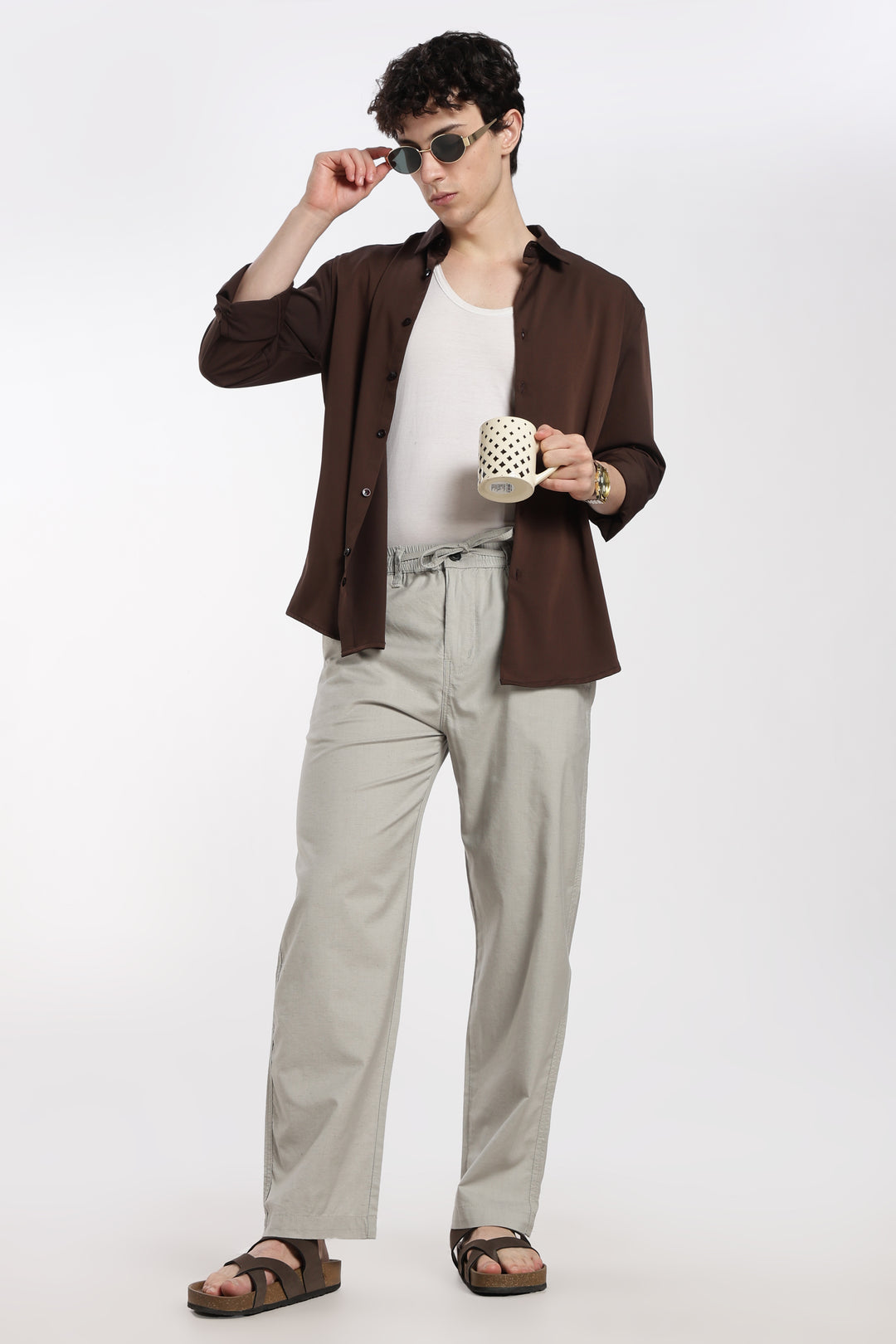 Man wearing a brown shirt, white tank top, and grey linen pants holding a mug on a white background