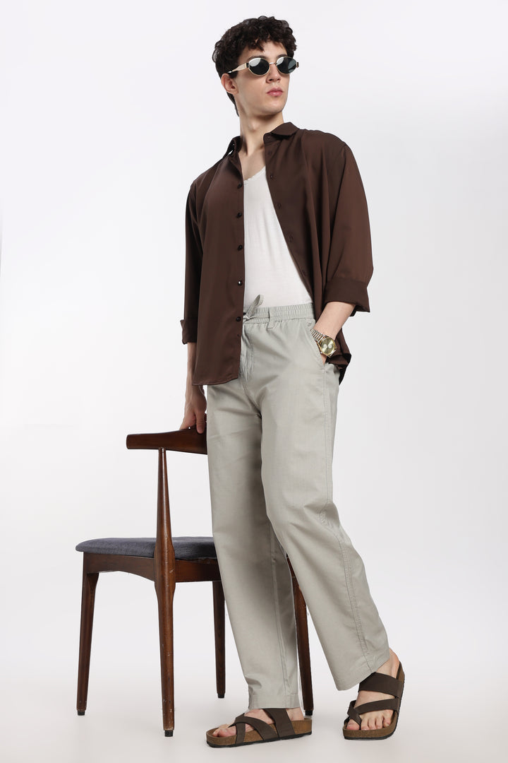 man wearing a brown shirt and grey linen pants standing next to a wooden chair on a white background