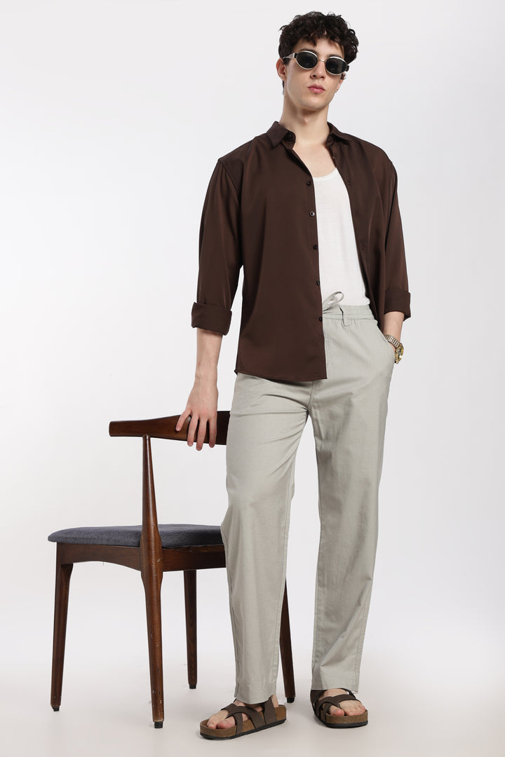 Man wearing a brown shirt and grey linen pants standing next to a wooden chair on a white background