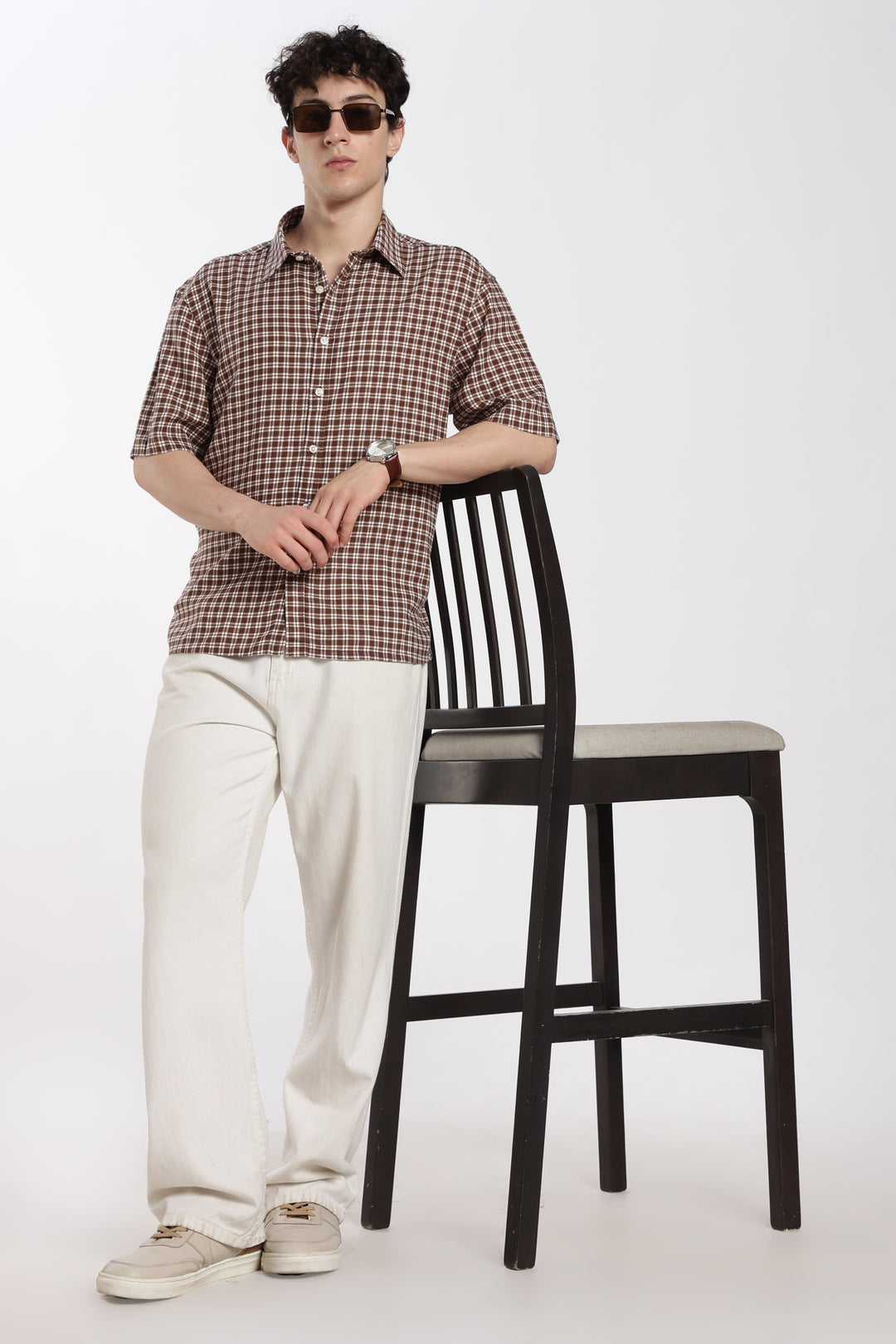 Man wearing a checkered half sleeve shirt and sunglasses standing next to a black chair on a white background