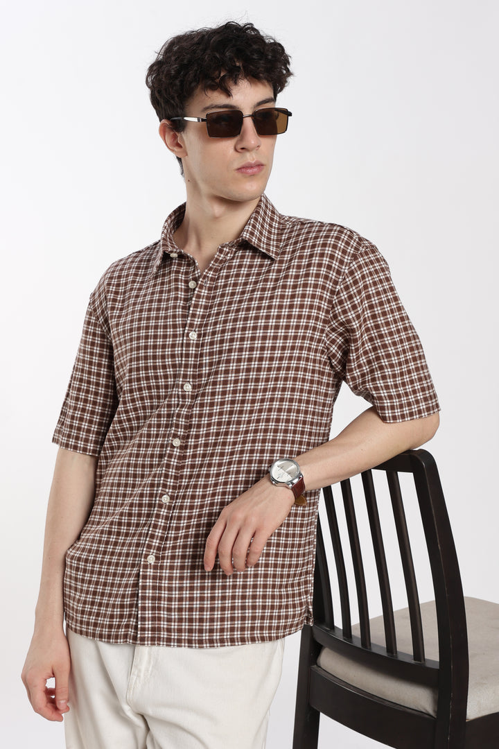 Man wearing a brown checkered half sleeve shirt and sunglasses, standing next to a chair on a white background