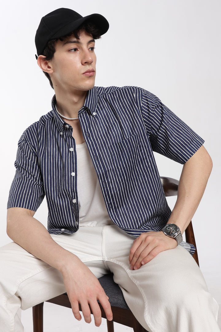 Man wearing a striped half sleeve shirt and cap sitting on a chair against a white background
