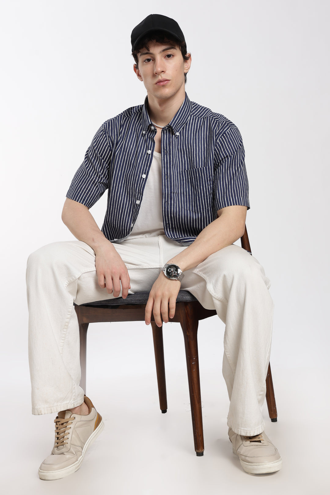 man sitting on a chair wearing a striped half sleeve shirt, white pants, and a cap on a white background