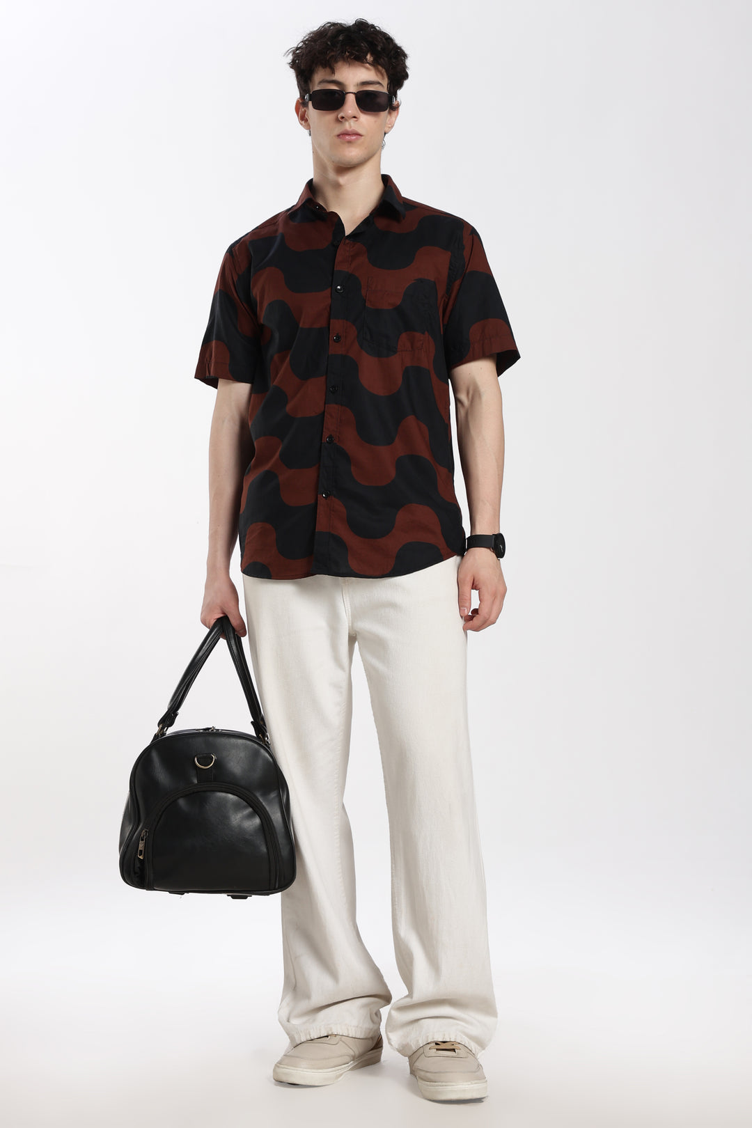 Man wearing a patterned half sleeve shirt and white pants, holding a black bag on a white background
