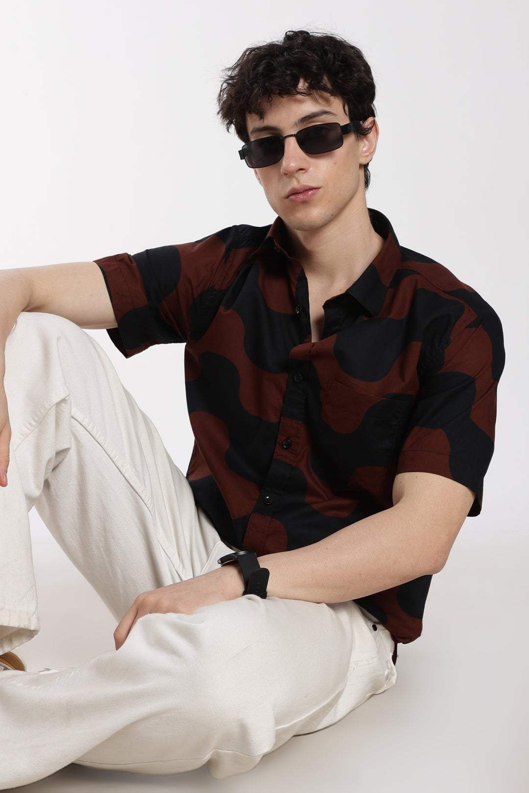 man wearing a patterned half sleeve shirt and sunglasses sitting on a white background