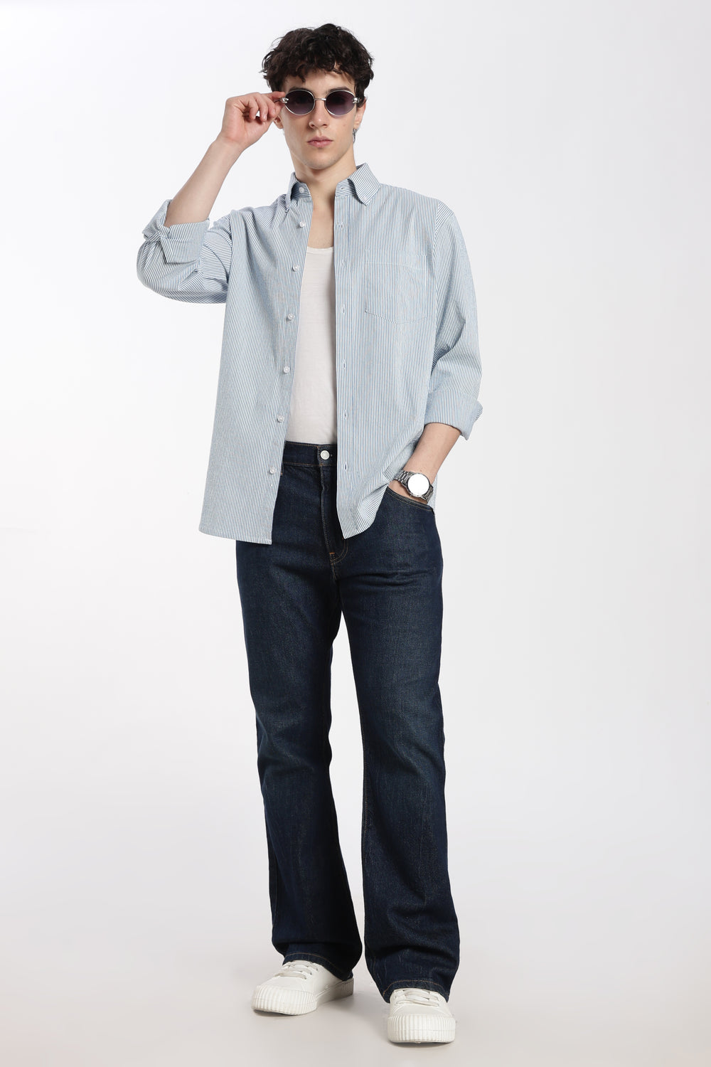 man wearing a blue stripe linen shirt, dark jeans, and white sneakers on a white background