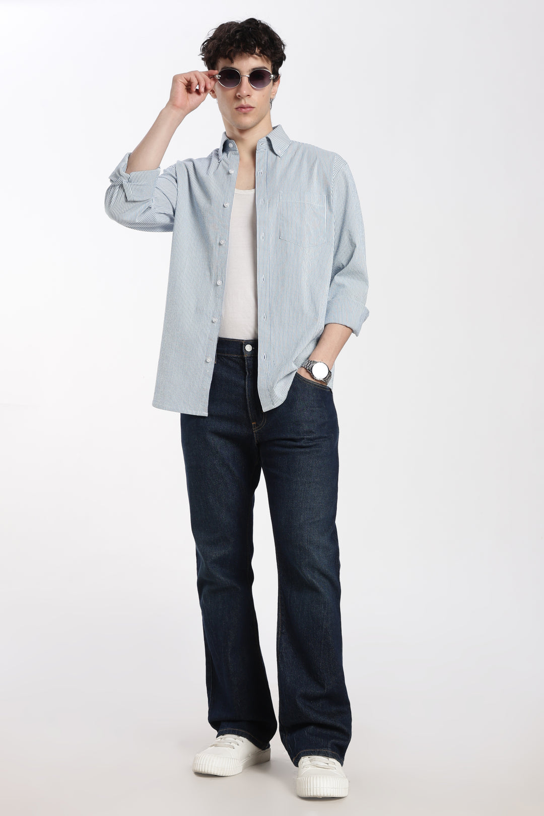 man wearing a blue stripe linen shirt, dark jeans, and white sneakers on a white background