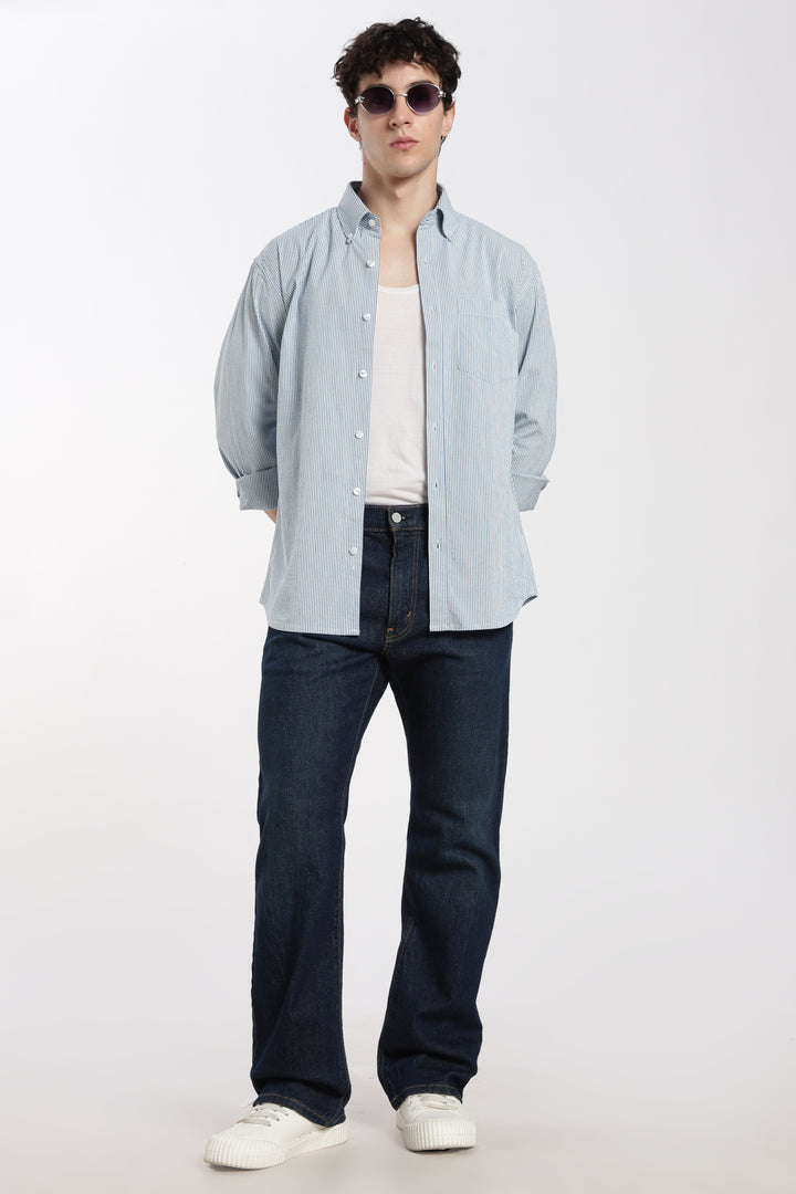 Man wearing a blue striped linen shirt, dark jeans, and white sneakers on a white background