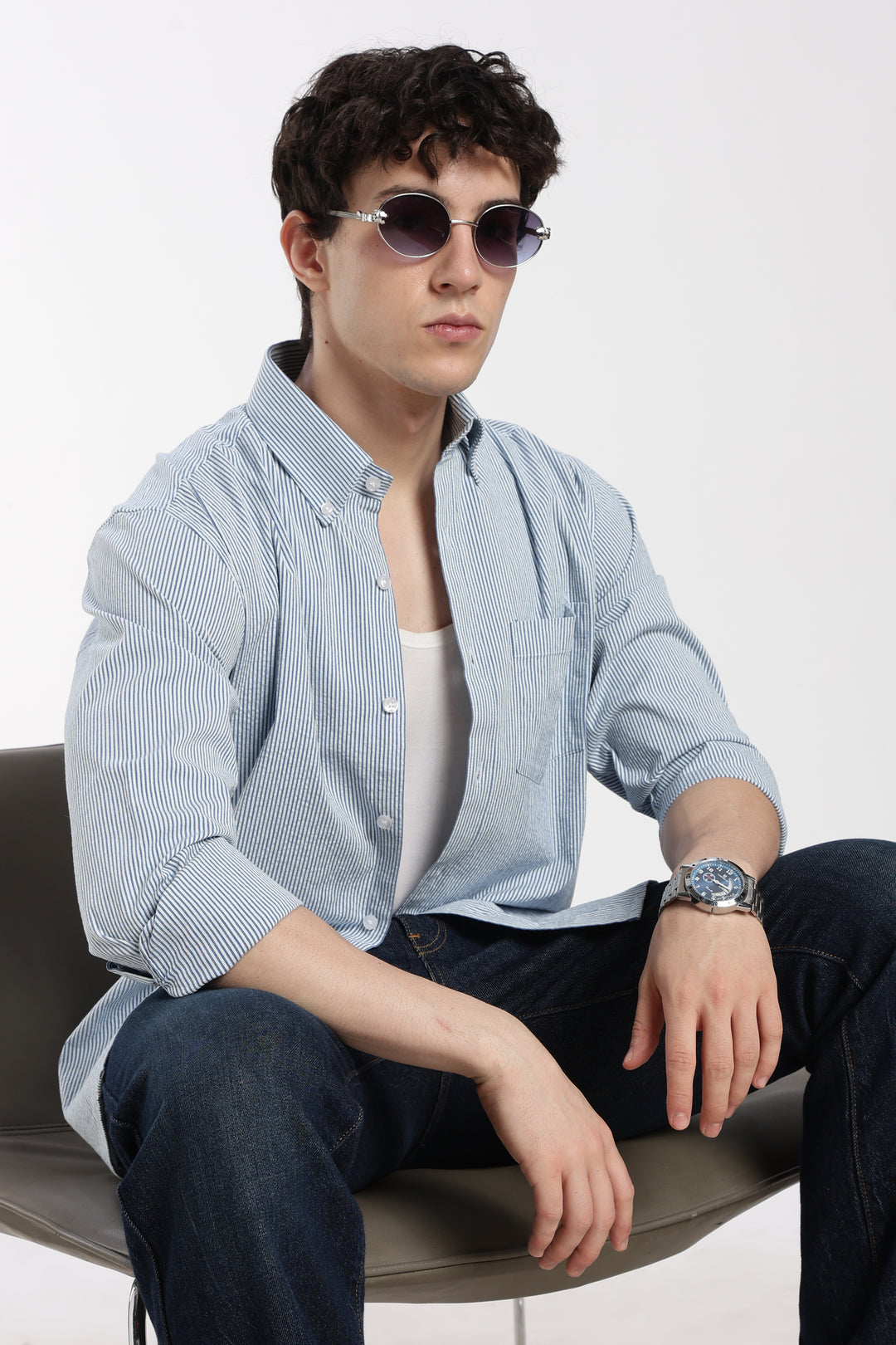 Man wearing sunglasses, a blue striped linen shirt, and dark jeans sitting on a chair.