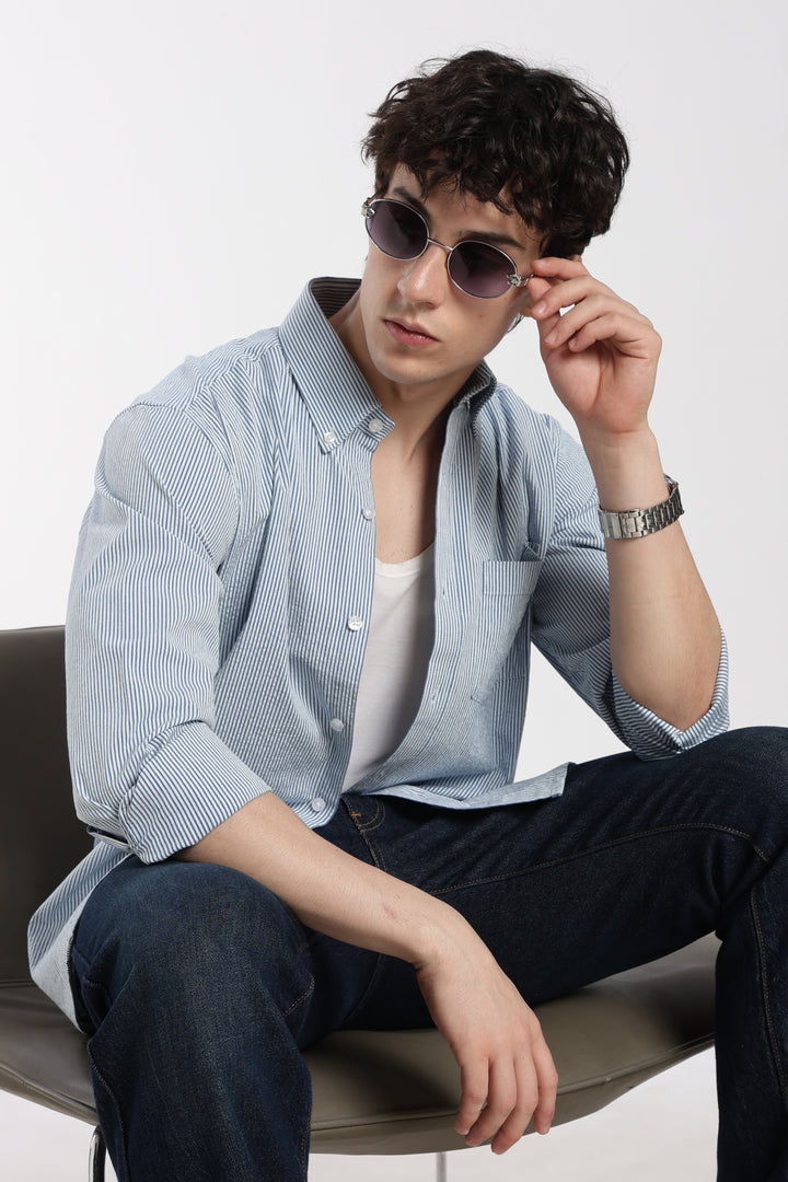 Man wearing sunglasses and a blue striped linen shirt sitting on a chair against a white background
