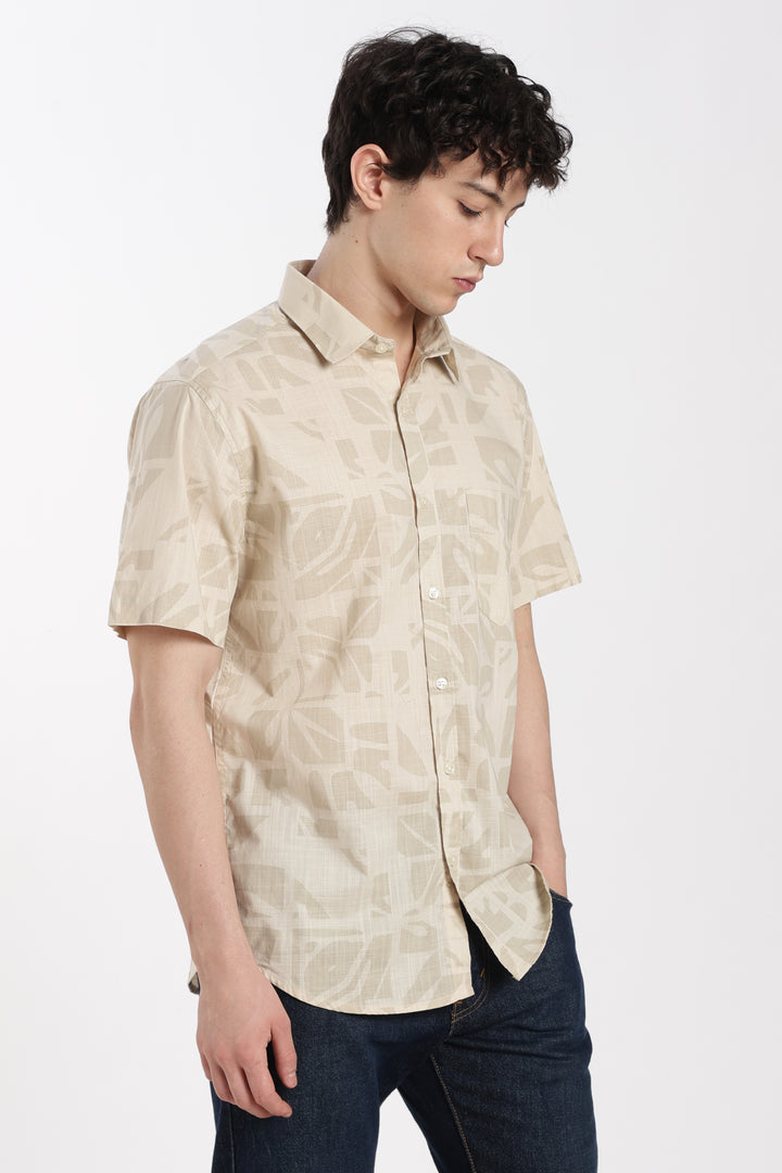 Man wearing a beige patterned half sleeve shirt and dark jeans on a white background