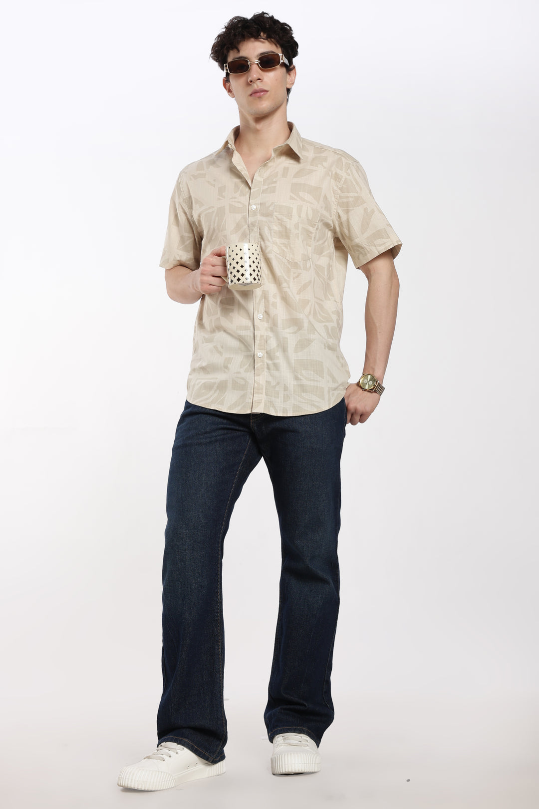 Man wearing a beige patterned half sleeve shirt and dark jeans, holding a polka dot cup against a white background