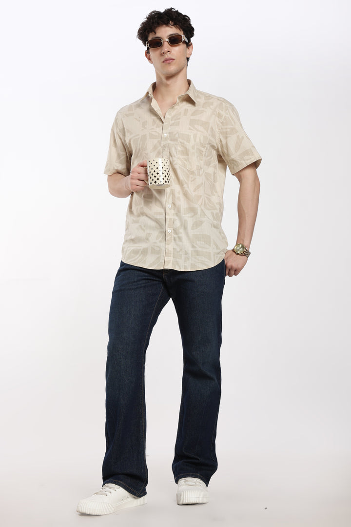 Man wearing a beige patterned half sleeve shirt and dark jeans, holding a polka dot cup against a white background