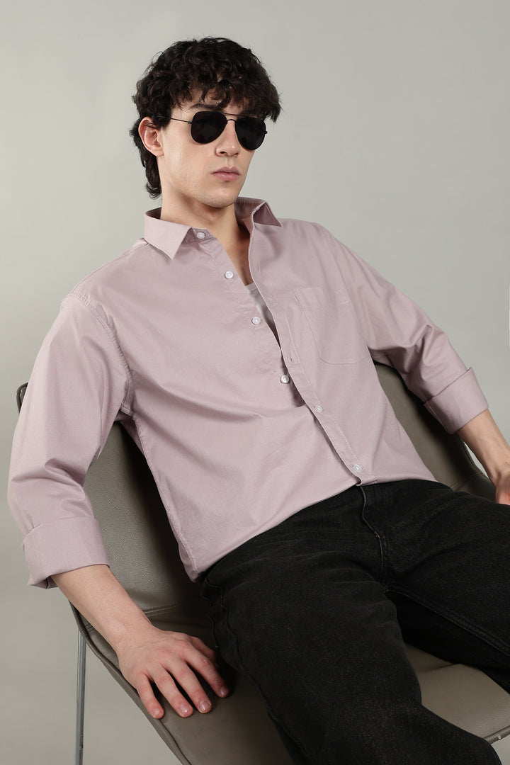 Man wearing a mute pink shirt and sunglasses sitting on a chair against a plain background