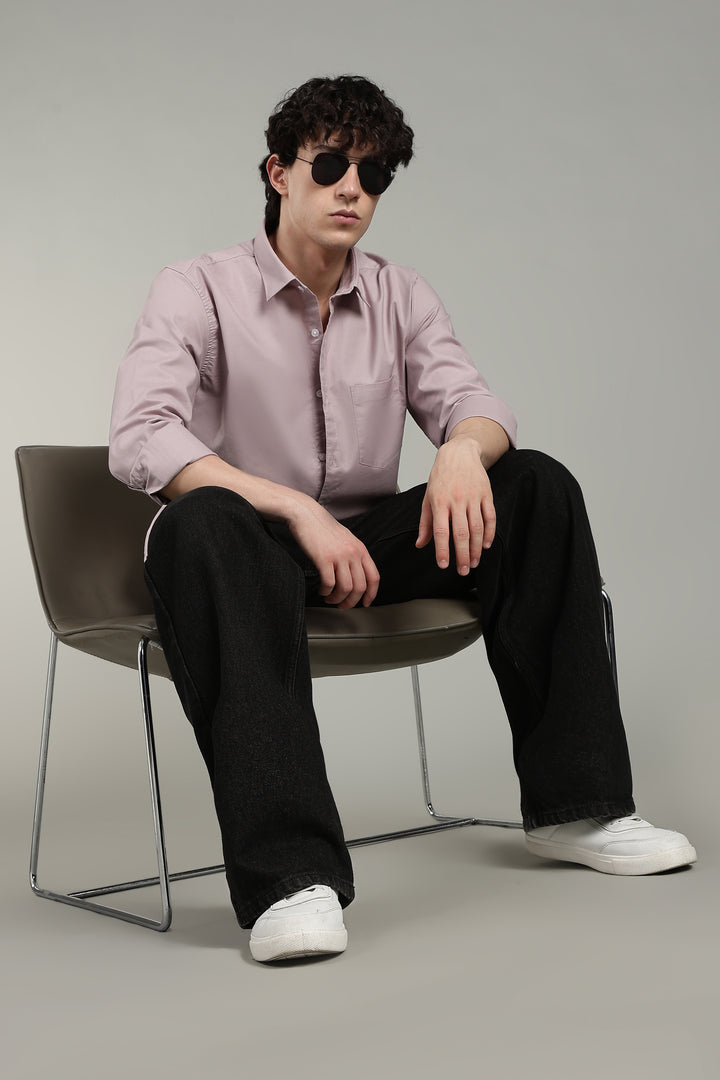 man sitting on a chair wearing a mute pink shirt, black pants, and sunglasses against a plain background