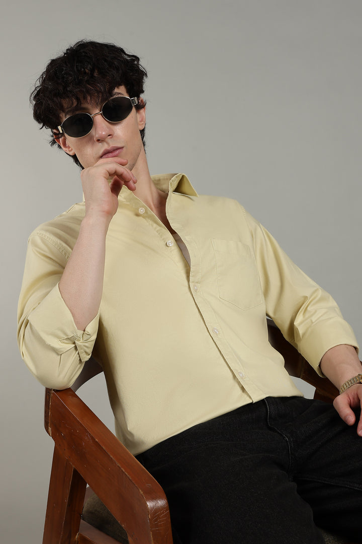 man wearing a light yellow shirt and sunglasses, sitting on a wooden chair against a plain background