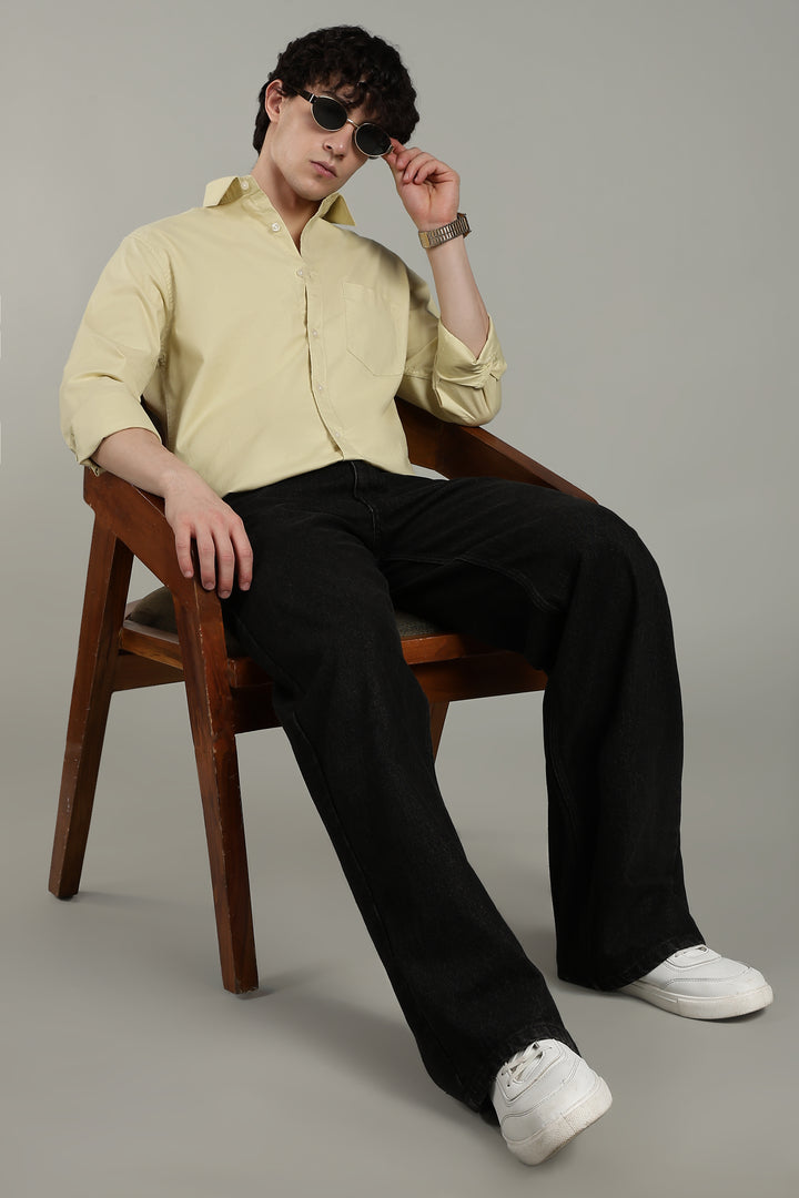 Man sitting on a wooden chair wearing a light yellow shirt, black pants, and white shoes.