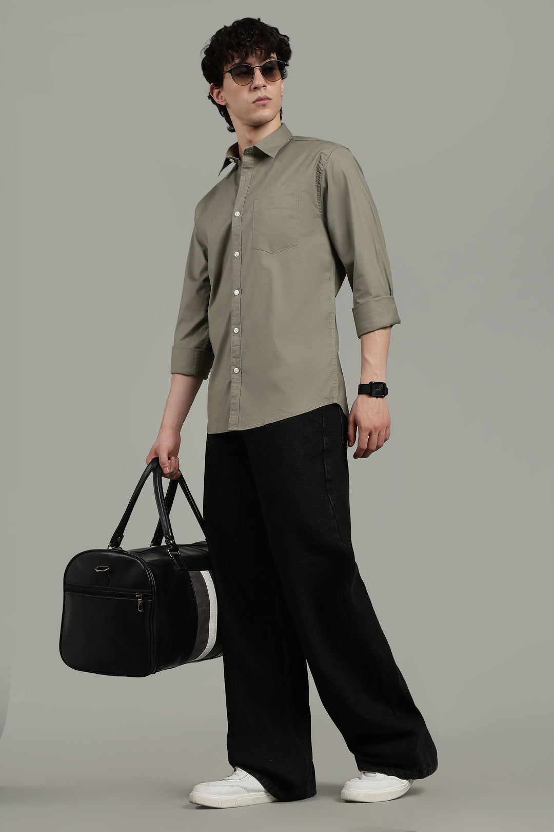 Man wearing a sage green shirt and black pants, holding a black bag on a gray background